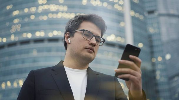 Man with earbuds standing with a phone on background of business centre - 写真・画像