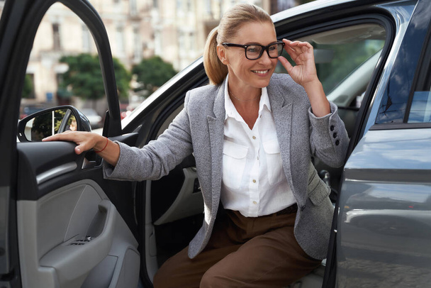 Porträt einer schönen und glücklichen Geschäftsfrau mit Brille, die aus ihrem modernen Auto steigt und lächelt. Anreise zum Schlachthof - Foto, Bild