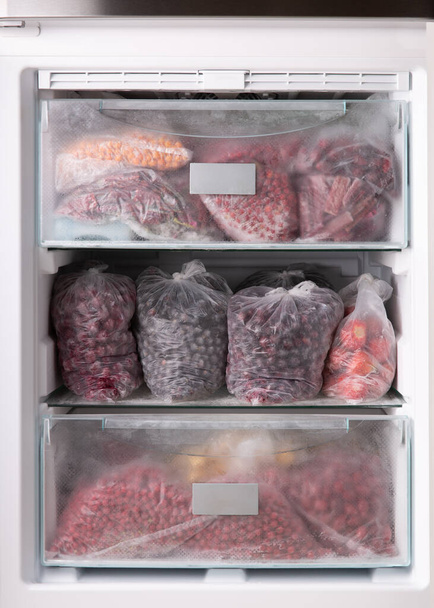 Frozen berries in transparent plastic bags in the freezer. Open refrigerator filled with frozen berries and fruits. Vegetarian food, healthy eating concept. - Photo, Image
