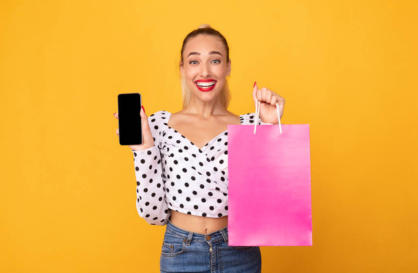 Happy woman showing black phone screen and shopping bag - Valokuva, kuva