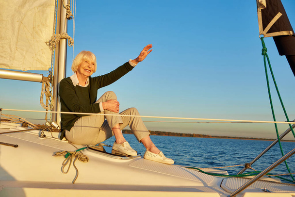 Šťastná starší žena sedící na boku plachetnice nebo jachty, plovoucí v klidném modrém moři, dívající se stranou a mávající na někoho - Fotografie, Obrázek