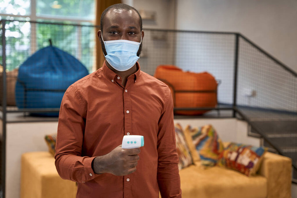 Ritratto di giovane impiegato africano che indossa una maschera protettiva medica con termometro a infrarossi per controllare la temperatura e guardare la macchina fotografica - Foto, immagini