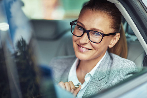 車の中でビジネスの女性。タクシーの後部座席に座ってカメラを見て笑顔で眼鏡をかけている陽気な中年女性 - 写真・画像