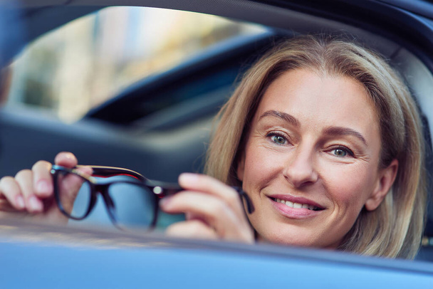 Portrait d'une femme d'affaires heureuse portant des lunettes et souriant à la caméra alors qu'elle était assise derrière le volant de sa voiture moderne, conduisant à travers la ville - Photo, image