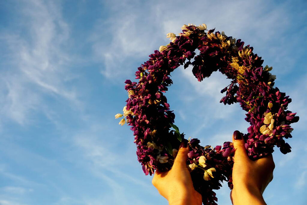 Στεφάνι από ροζ λουλούδια στο φόντο Blue Sky. Γυναικεία χέρια κρατώντας ένα στέμμα από μοβ λουλούδια σε ένα φόντο του μπλε ουρανού με λευκά σύννεφα. Χέρια με στεφάνι καλοκαιρινών λουλουδιών                         - Φωτογραφία, εικόνα