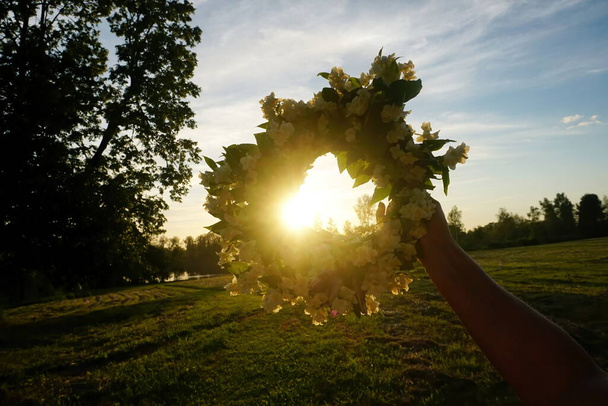夕方の夕日を背景に白いジャスミンの花のリースを保持する女性の手。美しい真夏の白花冠                      - 写真・画像