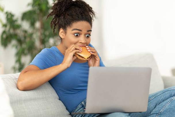 Afrikanisches Mädchen isst Burger mit Laptop auf Couch - Foto, Bild