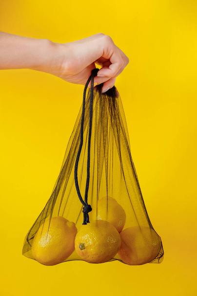 Mesh shopping bag with lemons on yellow background. - Фото, изображение