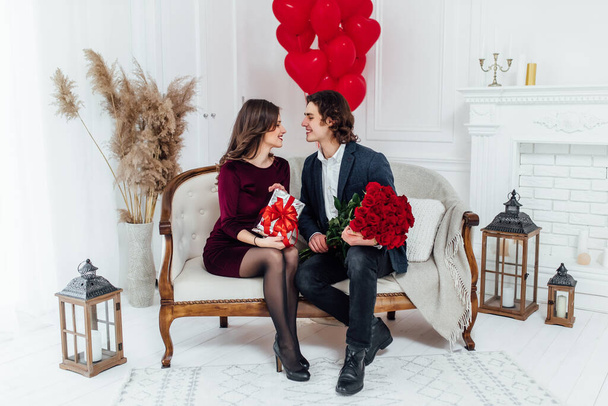happy couple sitting on the sofa and looking at each other with love, holding flowers and present in the hands with heart shaped balloons on the background - Zdjęcie, obraz