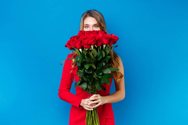 cute girl covering face with bouquet of red roses on blue background - Foto, imagen