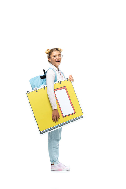 full length view of excited schoolgirl in eyeglasses holding paper cut copy book while looking at camera on white - Photo, Image