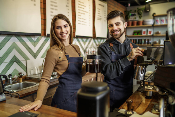 Couple of young smiling and pretty baristas at work. Professional barista team brewing coffee using coffee machine in coffee shop. - Φωτογραφία, εικόνα