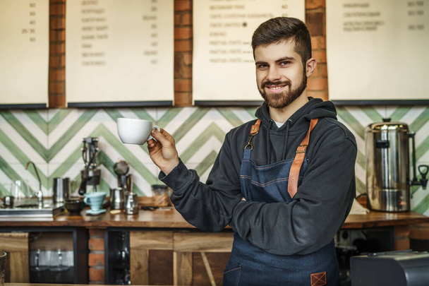 Portrait of  happy barista standing at trendy coffee shop, holding cup of coffe and smiling. - Photo, image