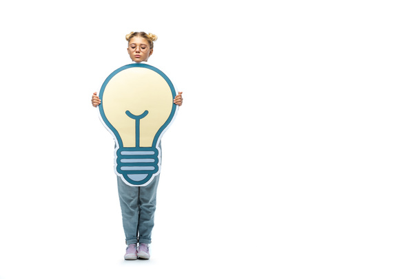 full length view of schoolgirl in jeans and eyeglasses holding paper cut light bulb on white - Фото, зображення