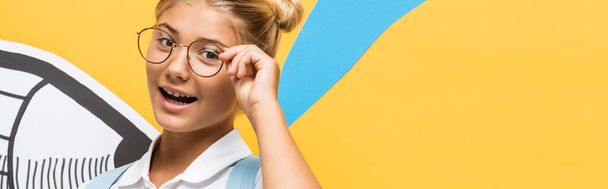 panoramic concept of joyful schoolgirl in eyeglasses looking at camera near paper cut pencil on blue and yellow - Photo, image