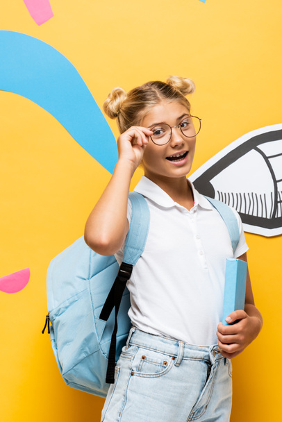 excited schoolchild touching eyeglasses and looking at camera while holding book near colorful elements and paper pencil on yellow - Foto, afbeelding