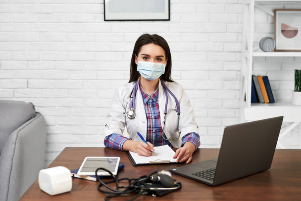 Portrait of a female doctor in her office in a protective mask. Woman at workplace in hospital with documents, laptop, tablet and tonometer. - Foto, imagen