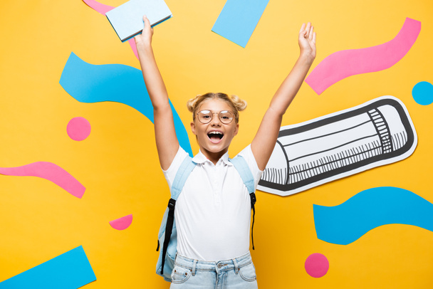 excited schoolgirl in eyeglasses screaming while holding book in raised hands near paper pencil and decorative elements on yellow - Fotó, kép