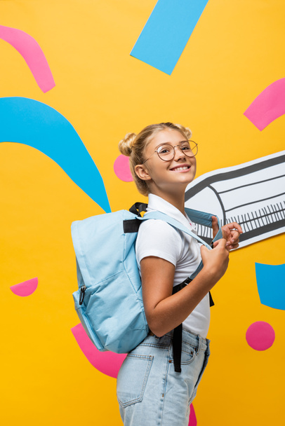 joyful schoolgirl with backpack looking away near paper pencil and colorful elements on yellow - Foto, immagini