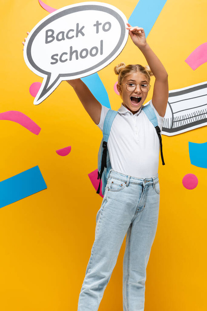 aufgeregtes Schulmädchen schreit, während es eine Sprechblase mit dem Rücken zur Schule in der Nähe von Papierkunst und Bleistift auf gelb hält - Foto, Bild