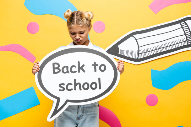 displeased schoolgirl holding speech bubble with back to school inscription near paper pencil and decorative elements on yellow  - Foto, immagini