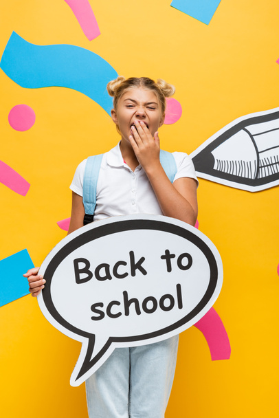 bored schoolgirl yawning while holding speech bubble with back to school lettering near paper pencil and colorful elements on yellow  - Photo, Image