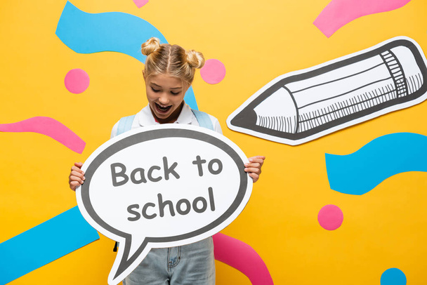 joyful schoolgirl holding speech bubble with back to school lettering near paper pencil and colorful elements on yellow - Foto, afbeelding