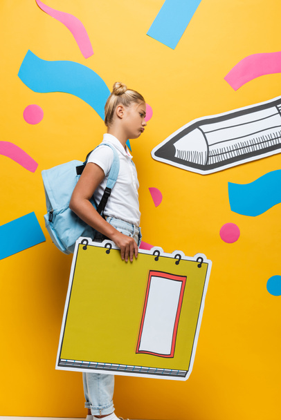zijaanzicht van boos schoolmeisje met notebook maquette op gele achtergrond met papier potlood en decoratieve elementen - Foto, afbeelding