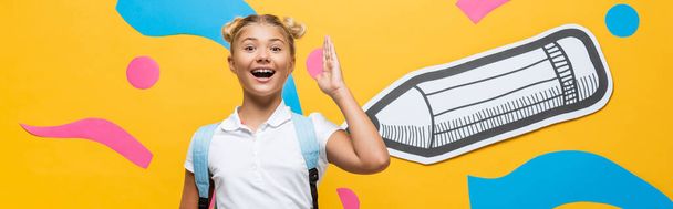 Website-Header des aufgeregten Schulmädchens hebt die Hand und schaut in die Kamera auf gelbem Hintergrund mit buntem Papier-Kunstwerk - Foto, Bild