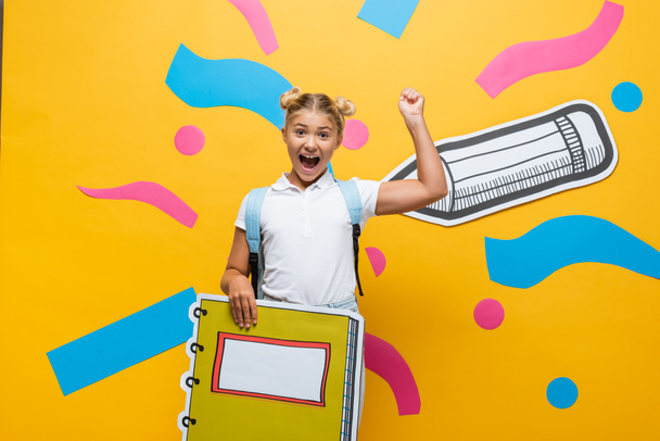 excited pupil with copy book maquette showing winner gesture and screaming on yellow background with decorative elements and paper cut pencil - Foto, Imagem