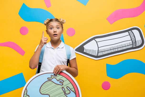 изумленный школьник с глобусным макетом, показывающим жест идеи и смотрящим в камеру рядом с бумажным карандашом и абстрактными элементами на желтом - Фото, изображение