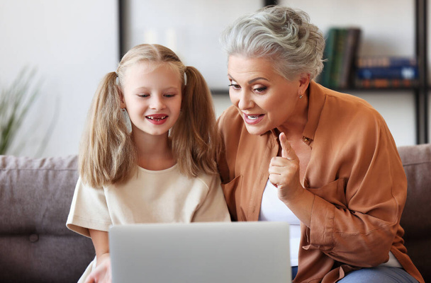Ältere Frau schüttelt den Finger und erklärt lächelnden Mädchen Daten, während sie auf dem Sofa sitzt und den Online-Kurs auf dem Laptop bei hom verfolgt - Foto, Bild