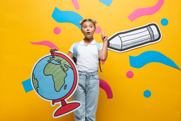 amazed schoolgirl showing idea gesture while holding globe maquette near paper pencil and colorful elements on yellow - Foto, Imagem
