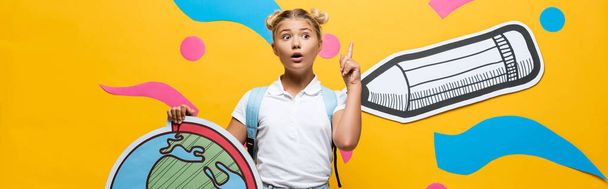 website header of excited schoolgirl showing idea gesture while holding globe maquette near colorful paper art on yellow - Photo, Image