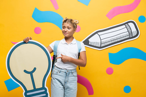 Confused schoolgirl pointing at paper light bulb near paper art on yellow background - 写真・画像