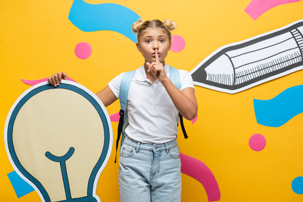Schoolgirl showing quiet gesture while holding decorative light bulb near paper art on yellow background - Foto, Imagen