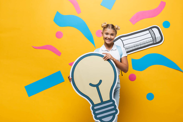 Schoolkid looking at camera while holding paper light bulb near paper art on yellow background - 写真・画像