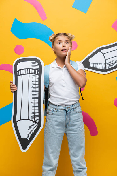 Worried schoolchild with backpack and paper pencil looking at camera on yellow background - Foto, Bild
