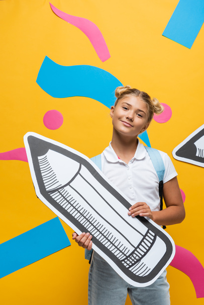 Schoolkid looking at camera while holding paper pencil on yellow background - Valokuva, kuva