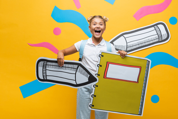 Opgewonden schoolkind met papier potlood en notebook op gele achtergrond - Foto, afbeelding