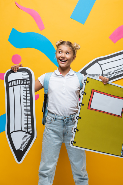 Schoolmeisje met rugzak, papier potlood en notebook in de buurt van decoratieve elementen op gele achtergrond - Foto, afbeelding