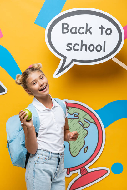 Schoolkind houdt appel in de buurt van spraakzeepbel met terug naar school belettering en papieren elementen op gele achtergrond - Foto, afbeelding