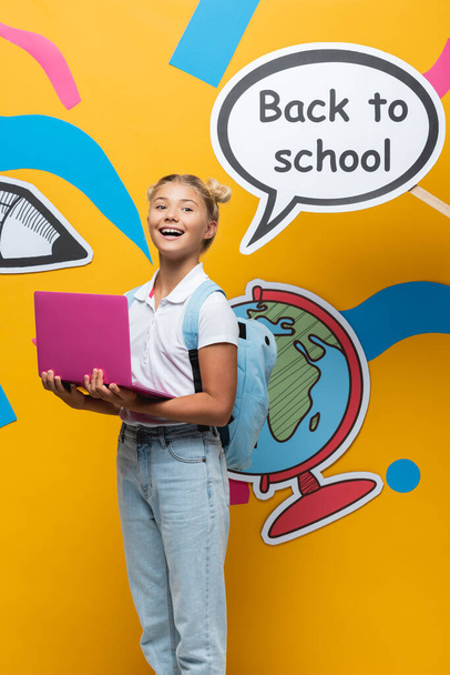 Liseli kız konuşma baloncuğunun yanında dizüstü bilgisayar tutuyor okula dönüş mektupları ve sarı arka planda kağıt resimleri var. - Fotoğraf, Görsel