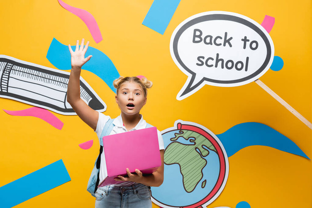 Opgewonden schoolmeisje met laptop zwaaiend naar camera in de buurt van spraakzeepbel met terug naar school belettering en papier kunst op gele achtergrond - Foto, afbeelding