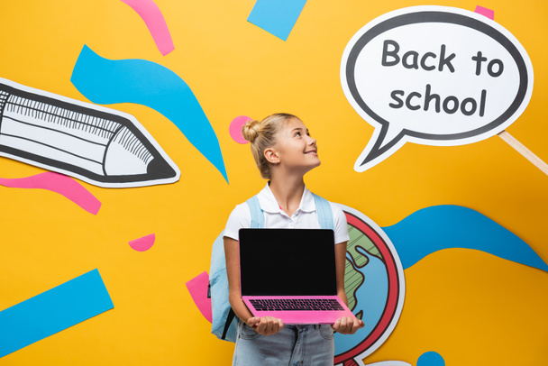 Sırt çantası ve dizüstü bilgisayarı olan liseli kız okula dönüş mektupları ve sarı arka planda kağıt resimleri olan konuşma baloncuklarına bakıyor. - Fotoğraf, Görsel