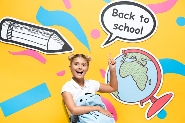 Schoolchild with backpack pointing at back to school lettering and paper elements on yellow background - Фото, изображение