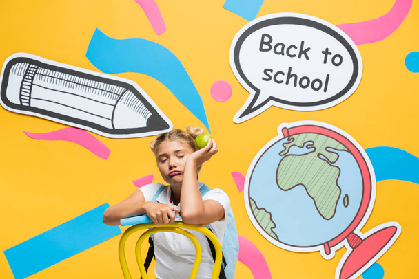 Tired schoolgirl with apple and book sitting near paper elements and speech bubble with back to school lettering on yellow background - Фото, изображение