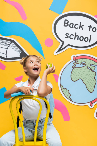 Ενθουσιασμένο παιδί με μήλο και βιβλίο κοιτάζοντας φούσκα ομιλία με πίσω στο σχολείο γράμματα και την τέχνη του χαρτιού σε κίτρινο φόντο - Φωτογραφία, εικόνα