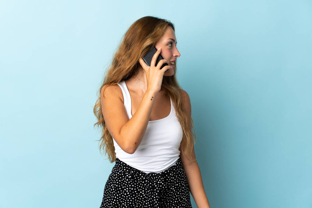 Νεαρή ξανθιά γυναίκα απομονωμένη σε μπλε φόντο κρατώντας μια συνομιλία με το κινητό τηλέφωνο με κάποιον - Φωτογραφία, εικόνα