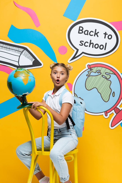 Excited schoolkid holding globe on chair near paper elements on yellow background - Foto, afbeelding
